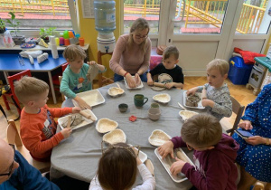 Grupa dzieci stoi przy stoliku wykonując prace ceramiczne.