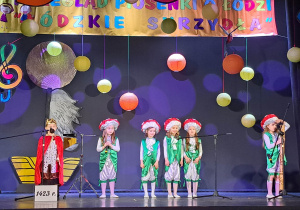 Grupa dzieci stoi na scenie, przed mikrofonami i śpiewa piosenkę o Łodzi.