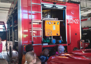 Dzieci stoją obok auta Straży Pożarnej i słuchają opowiadań Strażaka.