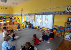 Dzieci siedzą na dywanie przed tablicą interaktywną i uczą się języka migowego.