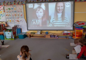 Dzieci siedzą na dywanie przed tablicą interaktywną i uczą się języka migowego.