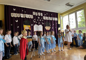 Dzieci stoją w sali, uczestniczą w uroczystości zakończenia roku przedszkolnego. Śpiewają piosenki, tańczą i recytują wiersze.