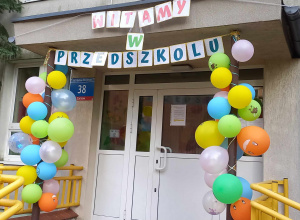 Wejście do przedszkola ozdobione kolorowymi balonami i napisem "Witamy w przedszkolu".