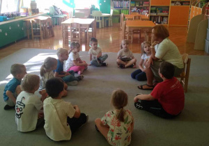 Dzieci z grupy Liski siedzą na dywanie w kole, słuchają głośno czytającej bajki nauczycielki.