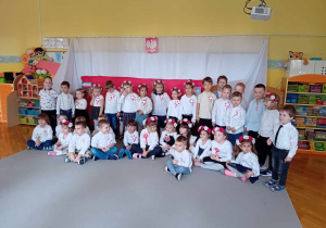 Dzieci stoją i siedzą, ubrane na galowo, na tle flagi Polski i godła Polski.