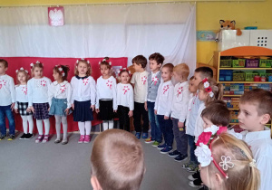 Dzieci stoją w kole, ubrane na galowo, śpiewają hymn Polski