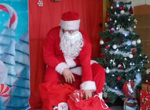 Mikołaj siedzi na krześle obok ozdobionej świątecznie choinki.