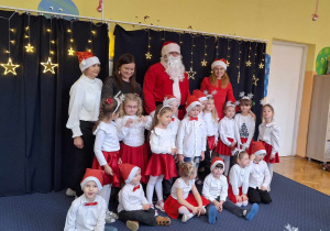 Dzieci z grupy Biedronki stoją z wychowawcami i Mikołajem na tle świątecznej dekoracji.