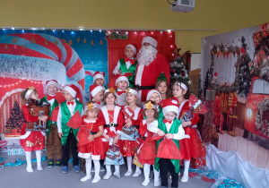 Dzieci z grupy Liski stoją z Mikołajem na tle dekoracji świątecznej.