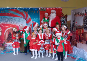 Dzieci z grupy Liski stoją z Mikołajem na tle dekoracji świątecznej.