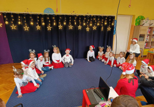 Dzieci z grupy Biedronki siedzą w kole na dywanie z opiekunem na tle dekoracji świątecznej.