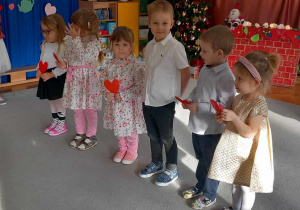 Dzieci stoją w półkolu, trzymając czerwone serca w rękach.