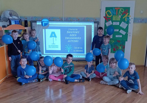 Dzieci z grupy Liski stoją i siedzą przed tablicą interaktywną z napisem "2 kwietnia Dzień Świadomości Autyzmu" z niebieskimi balonami.