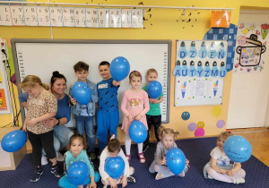 Dzieci z grupy Biedronki stoją i siedzą na dywanie z niebieskimi balonami.