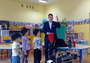 Iluzjonista w towarzystwie stojących obok dzieci wyjmuje z czerwonego woreczka skarpetkę.