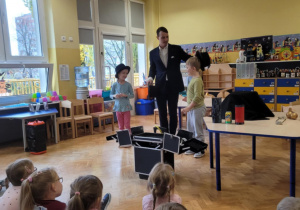 Iluzjonista w towarzystwie dziewczynki i chłopca, zdejmuje chłopcu czarny kapelusz, w którym była piłaczka.