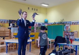 Iluzjonista obok, którego stoi chłopiec w czarnym kapeluszu, wyjmuje z kieszeni czarne kawałki materiału.