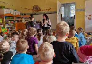 Dzieci siedzą na dywanie przed poetką Panią Urszulą Kowalską, która czyta dzieciom swoje utwory.