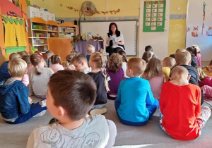 Dzieci siedzą na dywanie przed poetką Panią Urszulą Kowalską, która czyta dzieciom swoje utwory.