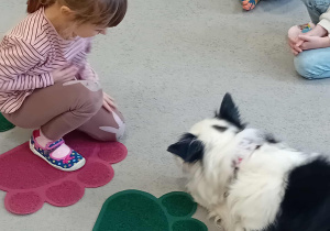 Sunia Lili leży na dywanie przed rozłożonymi sylwetami łapek psa, a obok siedzi dziewczynka.