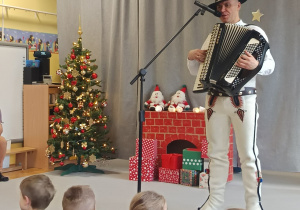 Góral Szczepan gra kolędy na akordeonie przed siedzącymi na dywanie dziećmi.