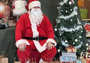 Mikołaj siedzi na krześle na tle świątecznej dekoracji, obok są prezenty.