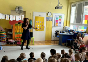 Pani Natalia stoi przed dziećmi siedzącymi przed nią i śpiewa piosenkę.