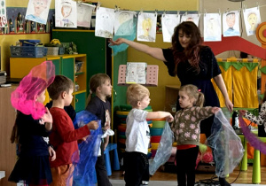 Pięcioro dzieci tańczy z kolorowymi chustami z panią Natalią na środku sali, przed nimi siedzą pozostałe dzieci.