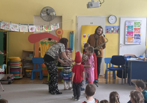 Dzieci stoją na środku sali, Pani Natalia przekazuje im grzechotki, opodal stoi mężczyzna trzymający saksofon.