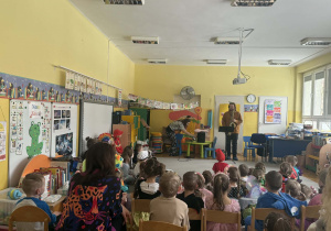 Mężczyzna stoi przed siedzącymi na dywanie dziećmi, grając na saksofonie.