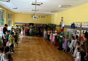 Dzieci, przebrane w wiosenne stroje tańczą w sali w kółku.