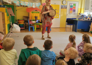 Dzieci siedzą na dywanie, słuchając Panią Nutkę, grającą na bębnie afrykańskim..