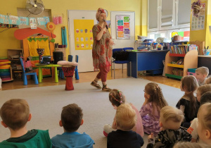 Dzieci siedzą na dywanie, słuchając Panią Nutkę, śpiewającą piosenkę.