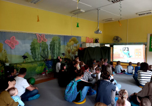 Dzieci wraz z rodzicami siedzą na dywanie w trakcie wiosennego seansu filmowego.