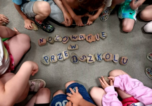 Dzieci siedzą na dywanie wokół ułożonego z kamieni napisu "Nocowanie w przedszkolu".