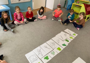 Realizacja Innowacji pedagogicznej "Słucham , Czuję, Analizuję Poezję - Wspieranie Rozwoju Małego Dziecka" - wiersz "Żabi koncert". Na dywanie rozłożone są ilustracje żab i instrumentów. Wokół siedzą dzieci.
