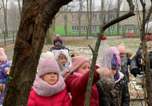 W ramach projektu "Jestem małym przyjacielem przyrody" - dzieci zebrane są wokół dziewczynki, która sypie ziarna dla ptaków do karmnika.