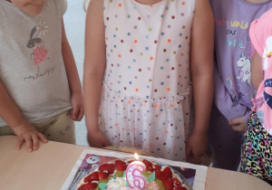 6 urodziny Mai. Dziewczynka stoi, a na stoliku jest tort. Maję otaczają dzieci.