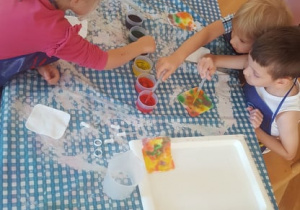 Kilkoro dzieci siedzi przy stoliku i maluje płatki kosmetyczne przy pomocy pipetek i wody zabarwionej barwnikami spożywczymi.