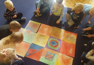 Dzieci siedzą na dywanie i podziwiają swoje prace ułożone w centralnym punkcie.